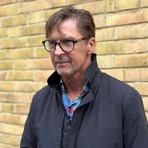 Per-Anders står framför en gul tegelvägg, han bär svarta glasögon och en rock.