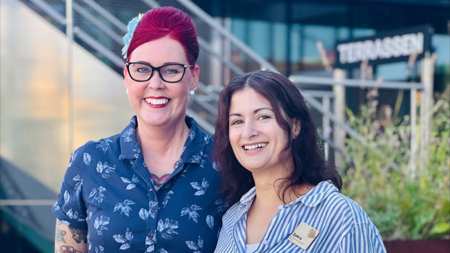Elin har sitt starkt röda hår uppsatt och bär en ljusblå blomma i det. Hon bär glasögon med svarta bågar och är klädd i en blåmönstrad skjorta. Zahra har mörkt långt hår och är klädd i en blå- och vitrandig skjorta. Båda ler mot kameran.
