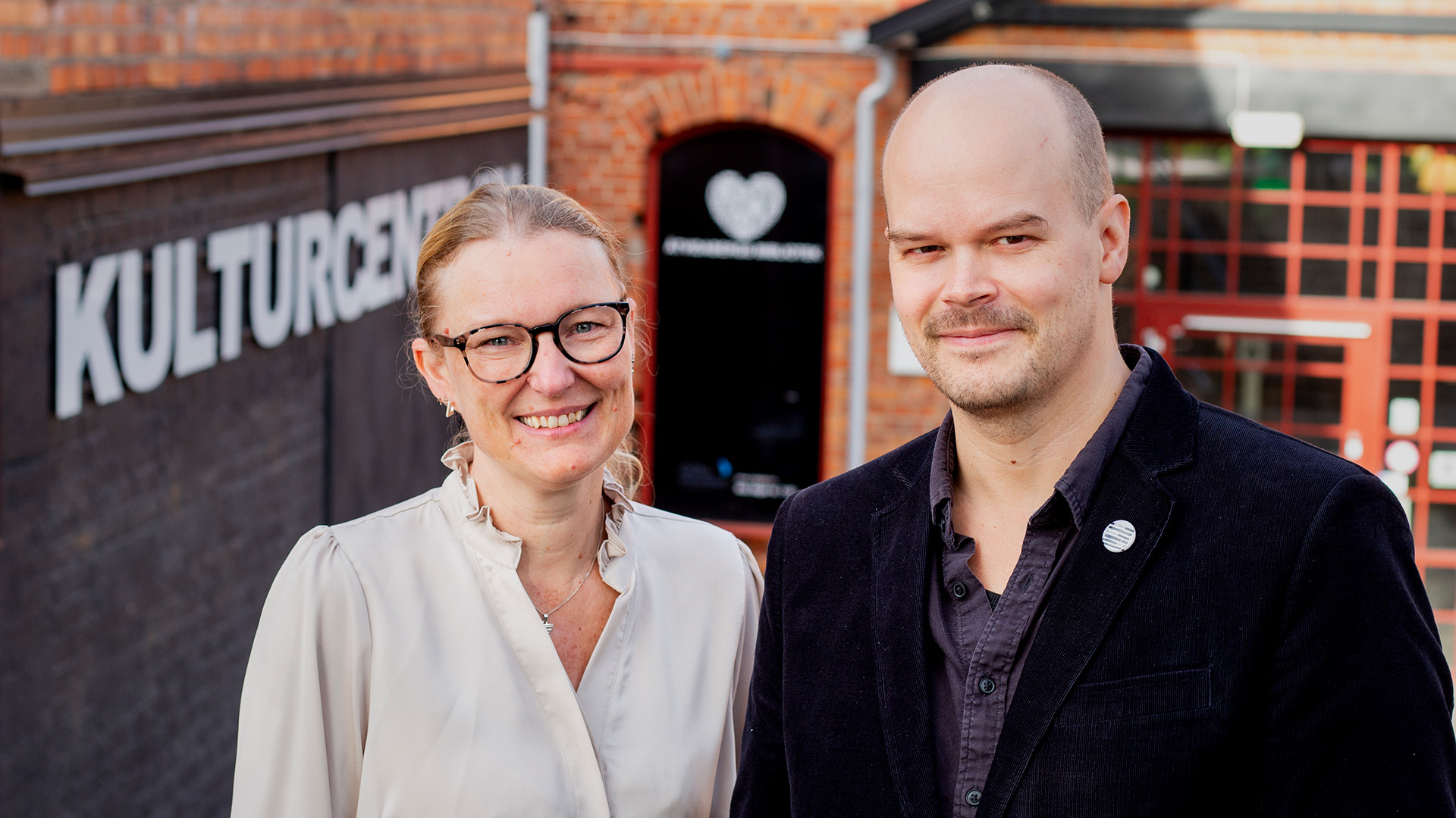 Anna Danielsson och Sebastian Laudon står bredvid varandra utomhus. I bakgrunden finns en byggnad i rött tegel och på väggen står det Kulturcentrum. Anna är blond och har håret i en hästsvans. Hon är klädd i en vit blus och bär glasögon. Sebastian har rakad hjässa, lite skäggstubb och är klädd i en mörk skjorta och en svart kavaj. Båda tittar mot fotografen och ler.