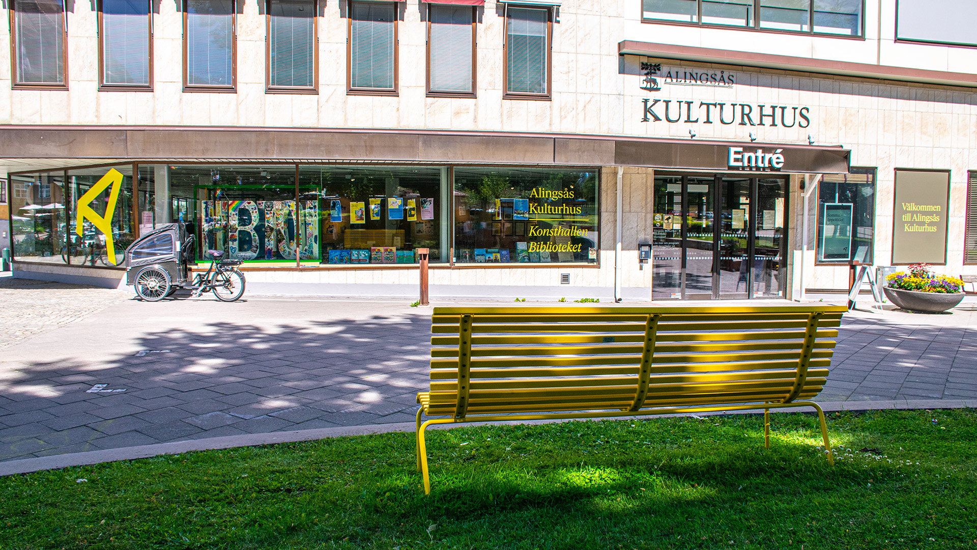 Kulturhuset i Alingsås, en byggnad med beige fasad och stora skyltfönster på första våningen. En lådcykel står parkerad utanför. En gul bänk är placerad i en park mitt emot entrén.