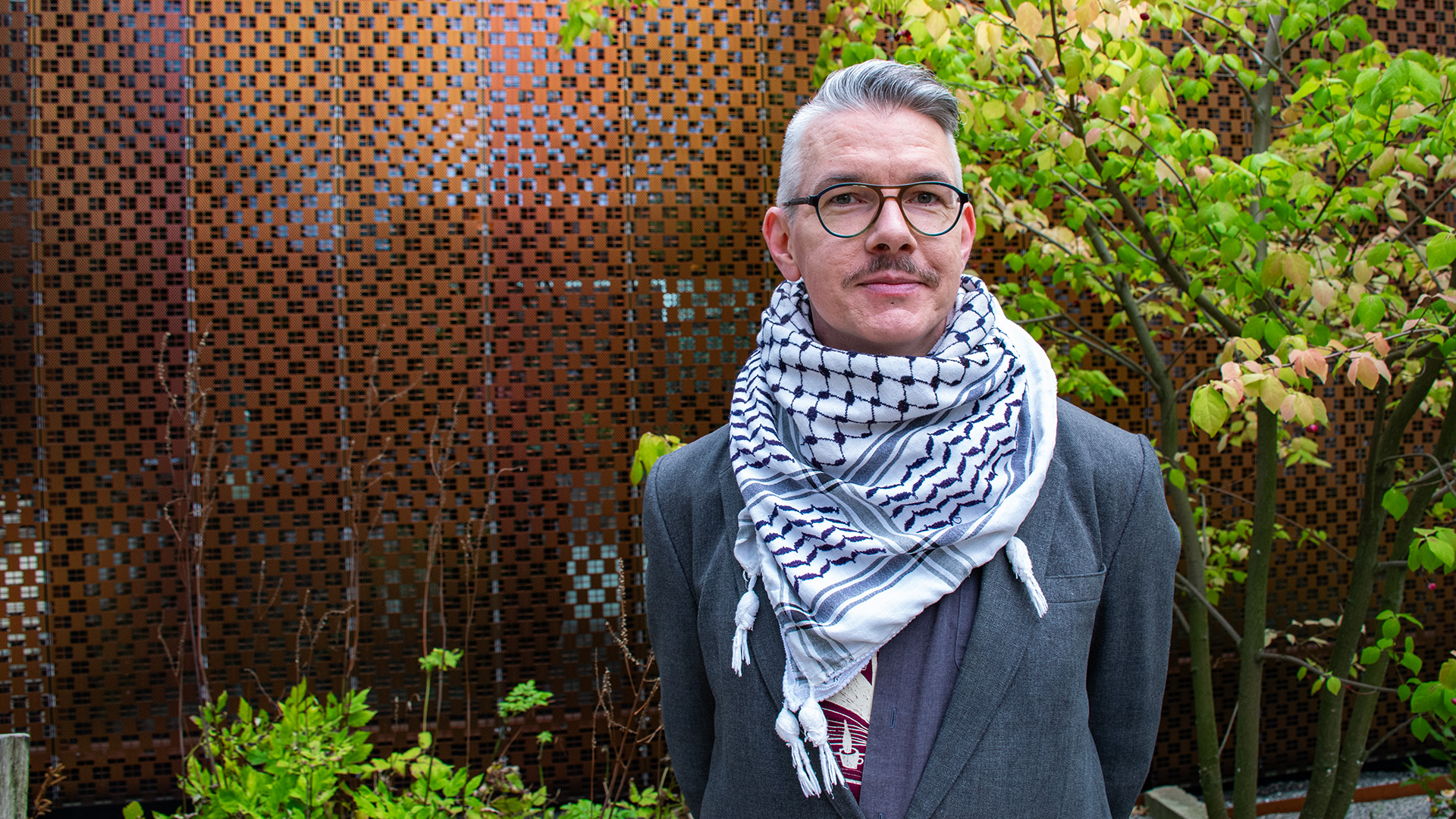 Olov Kriström är i 40-årsåldern. Han har gråsprängt kortklippt hår som är kammat bakåt, han är klädd i en grå kavaj och bär en palestinasjal runt halsen. Han har moderna glasögon och en mustasch. Bakom honom finns en kopparfärgad genombruten vägg och ett litet träd som håller på att få gula löv.