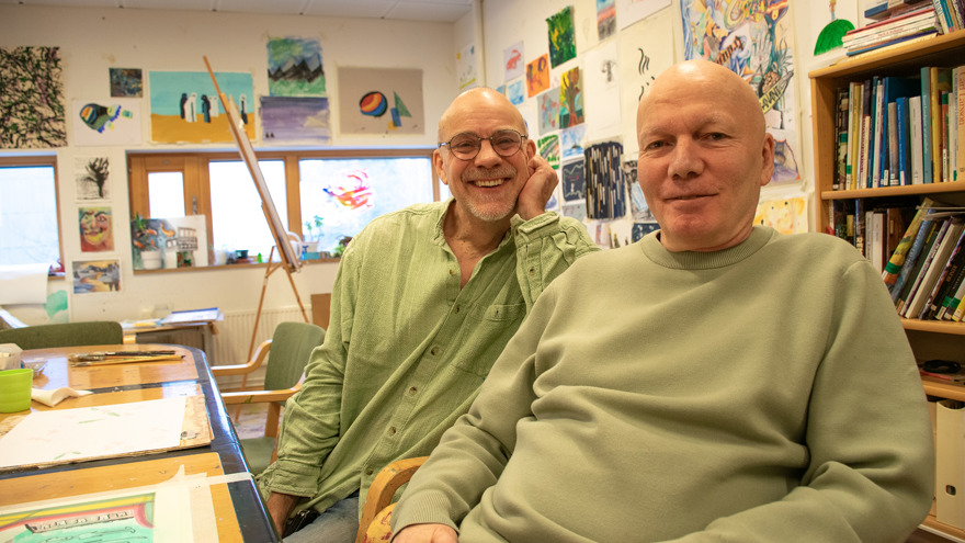 Henrik Wig och Stefan Karlsson sitter på varsin stol i en konstateljé. Runt dem finns målningar och teckningar uppsatta på väggarna. Henrik är klädd i en grön skjorta och har grått skägg och runda glasögon. Stefan har en grön tröja och har rakat huvud. Båda männen är i medelåldern.