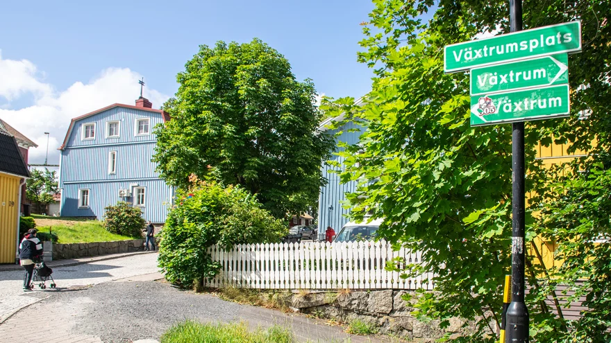 En gatubild från Lerum. En person med rollator går uppför en backe, runt om vägen ligger trähus i ljusblått och gult. I förgrunden finns gröna vägskyltar med texterna "Växtrum" och "Växtrumsplats". Solen skiner.
