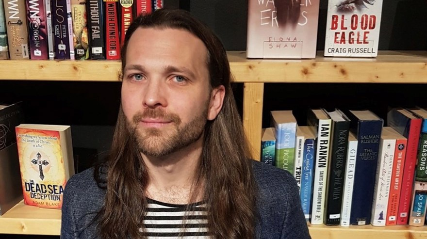 Pontus Kjellberg har långt brunt hår och skägg. Bakom honom finns en bokhylla fylld med böcker.