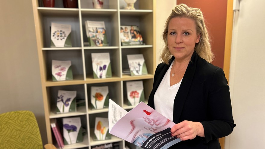 Blond kvinna i svart kavaj läser en rapport och  tittar in i kameran