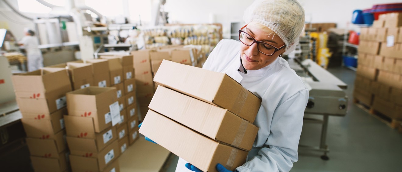 Halvbild på en person i vit rock och hårnät bär som på tre pappkartonger. Personen befinner sig i fabrik.