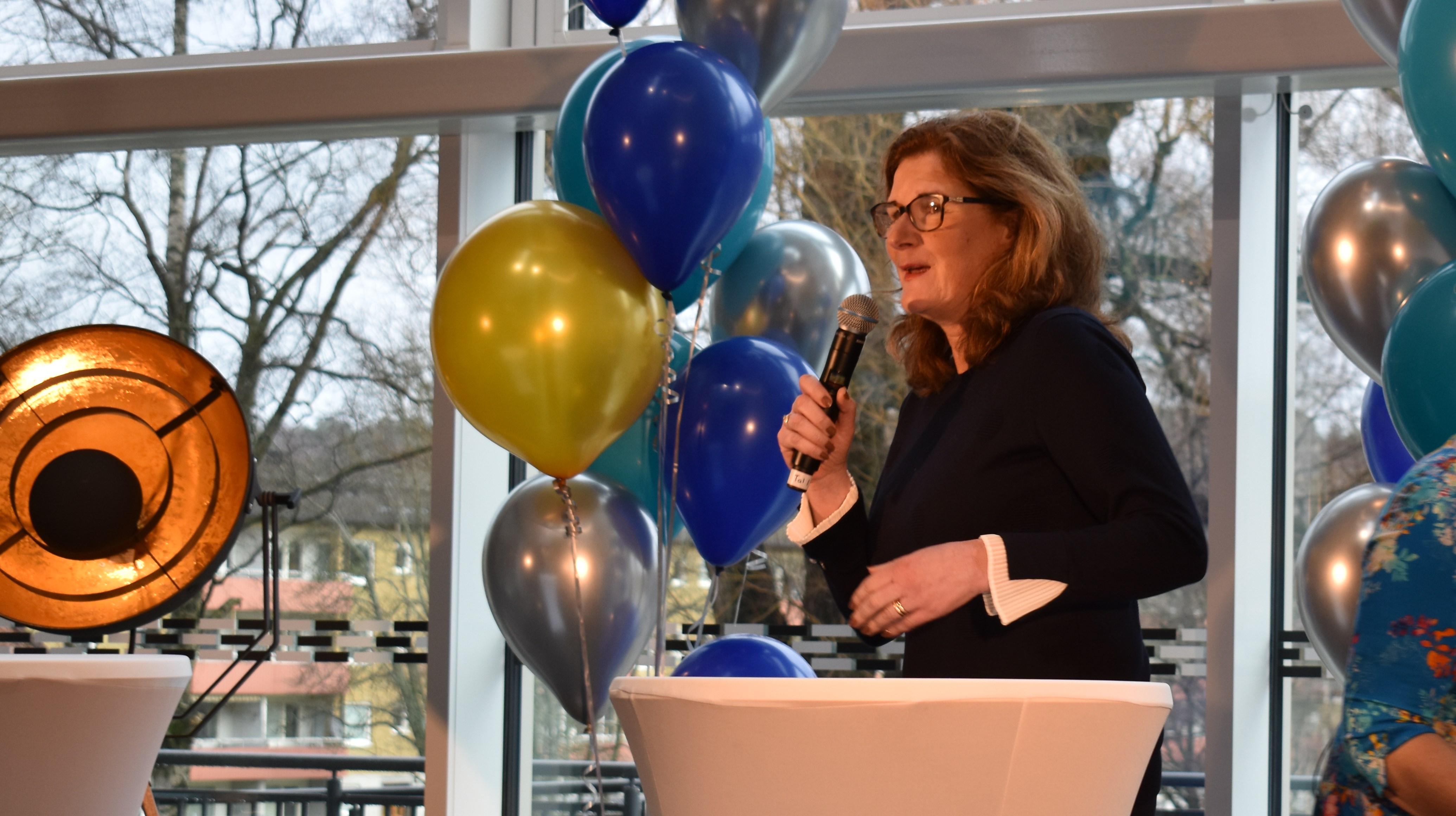 Kvinna i svart talar ien mikrofon. Ballonger i bakgrunden.