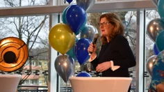 Kvinna i svart talar ien mikrofon. Ballonger i bakgrunden.