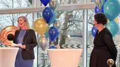 Två kvinnor står på en scen. Båda har mikrofonset på sig. Kvinnan till vänster talar.Bakom dem hänger ballonger.