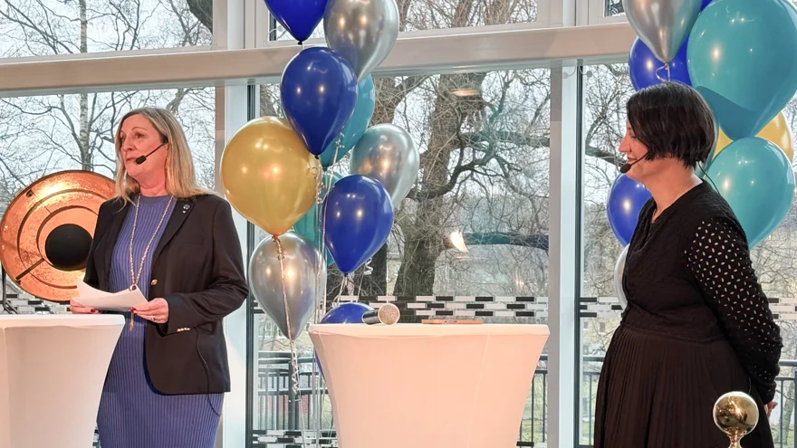 Två kvinnor står på en scen. Båda har mikrofonset på sig. Kvinnan till vänster talar.Bakom dem hänger ballonger.