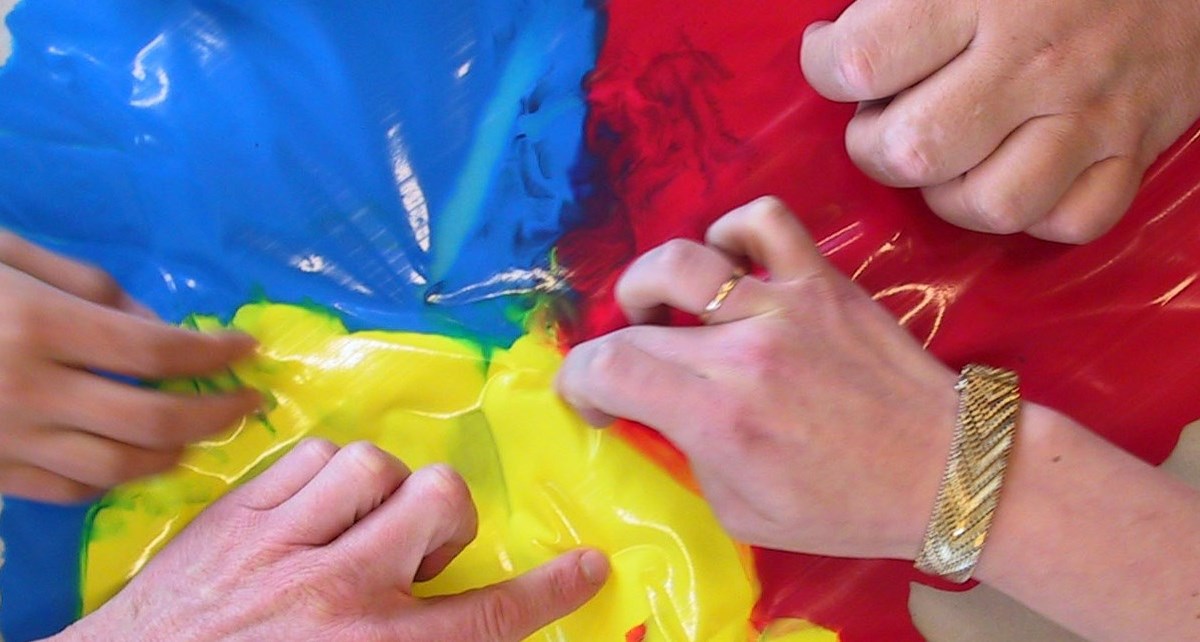 Flera händer trycker på plast som täcker över gul, röd och blå färg