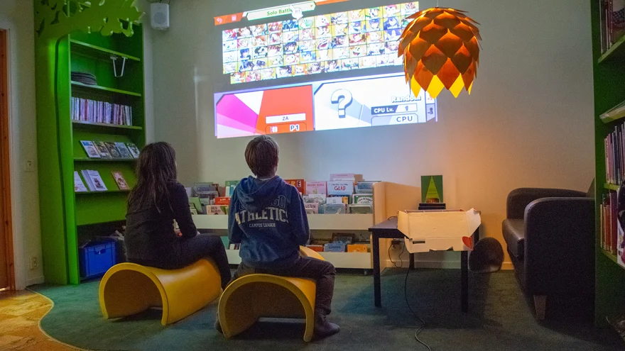Två barn sitter på varsin pall i ett mörkt rum. På väggen framför dem är ett tv-spel projicerat. De håller i varsin spelkontroll och har blicken fäst på spelet på väggen. Två gröna bokhyllor står utmed väggarna och ett tråg med barnböcker står under spelet på väggen. På golvet ligger en grön mjuk matta.