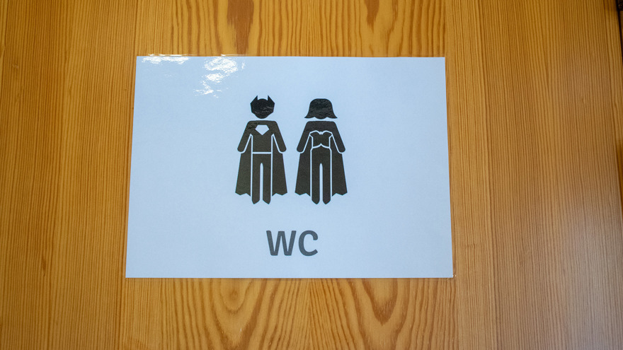 Skylt på toalettdörren på biblioteket i Kungshamn. På den vita skylten finns en manlig och en kvinnlig superhjälte. De bär båda mantlar och huvudbonader.