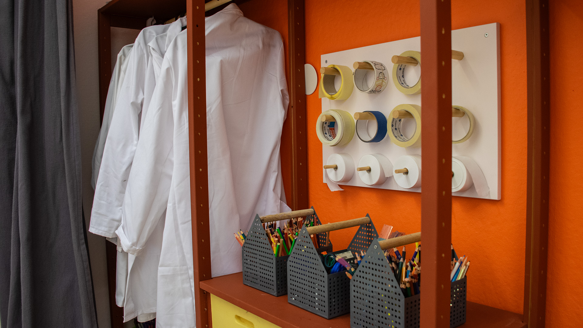 Vita labbrockar hänger på en stång. Tråg med färgglada pennor står på en hylla och stora rullar med tejp hänger på väggen. 