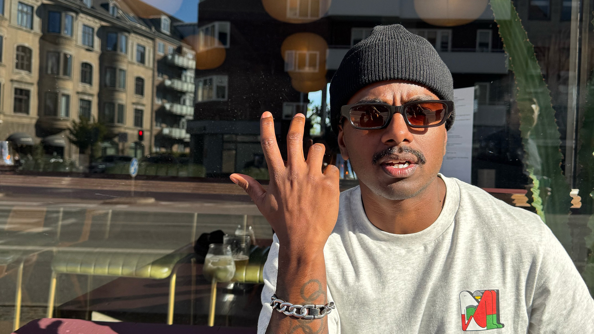 Loui Sand sitter utomhus i solen, kanske på en uteservering. Ett stort fönster bakom honom speglar omgivningen. Loui har en svart mustasch, är klädd i en grå t-shirt, bär en svart mössa och solglasögon. Han håller upp ena armen och han har tatueringar och ett brett silverarmband. Han är gissningsvis i 30-35-årsåldern.