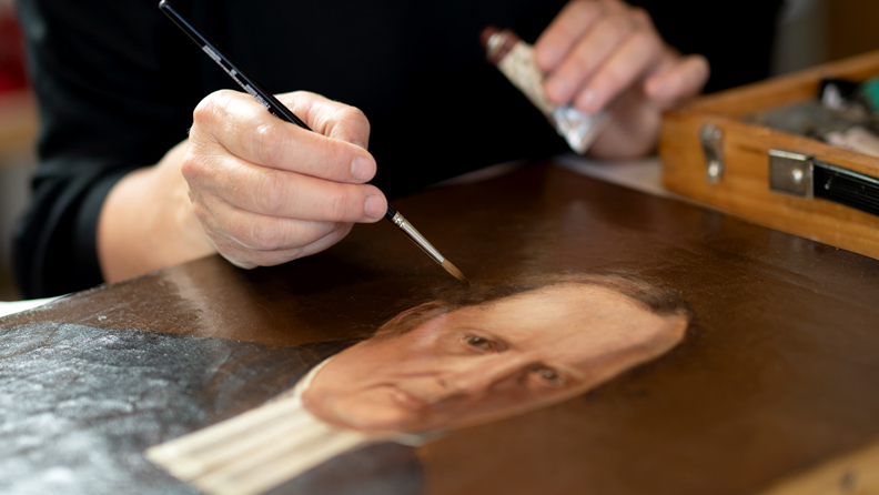 Vi blickar ner mot ett arbetsbord. På bordet ligger en målning, en målarlåda, färgtuber och burkar. Vi ser målerikonservatorn, en kvinna, rakt uppifrån. Hon håller i en palett med vänster hand och med höger hand retuscherar hon målningen på bordet med en mycket tunn pensel.