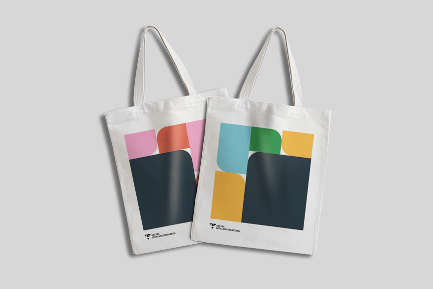 Två tygkassar mot grå bakgrund. På kassen är det grafiska former i form av fyrkanter med ett rundat hörn, VGR:s långa logotyp är längst ner till vänster. På den ena tygkassen är det grå huvudfärg på den största grafiska formen. De fyra mindre formerna är i rosa och orange komplementfärg. På den andra tygkassen är det också grå huvudfärg på den största formen. De fyra mindre formerna är består av två gula, en ljusblå och en grön.