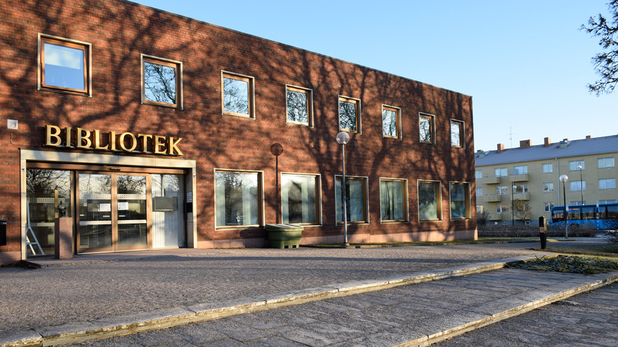 Falköpings bibliotek är en tvåvåningsbyggnad i rött tegel. Fönstren är mång och kvadratiska, ovanför glasdörrarna står det BIBLIOTEK med gula bokstäver. Biblioteket ligger i morgonsol, på vägen bredvid passerar en blå buss.