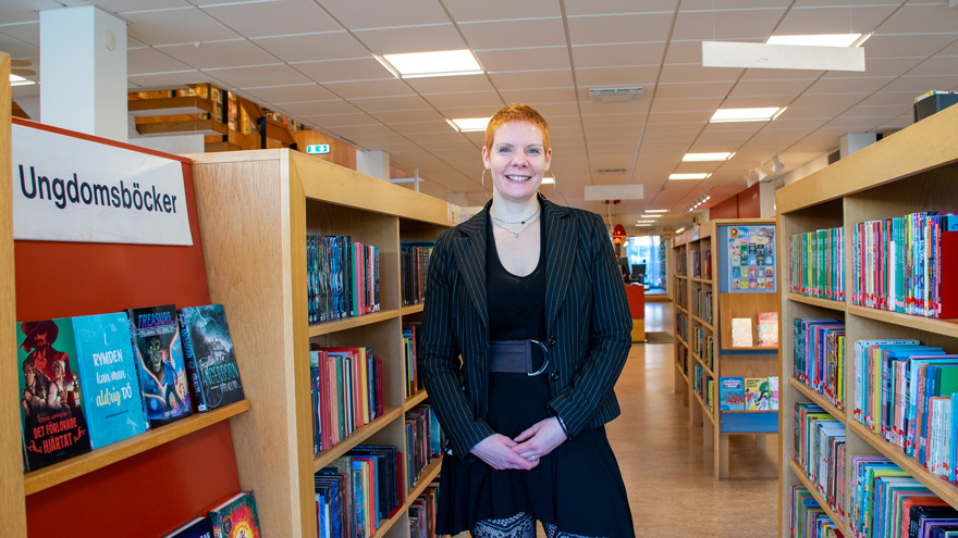 Kerstin Hellman är lång, har kortklippt rött hår och är klädd i svart klänning och en svart kavaj. Hon bär stora ringar i öronen och ler stort mot fotografen. Hon står mellan två bokhyllor på biblioteket i Falköping.