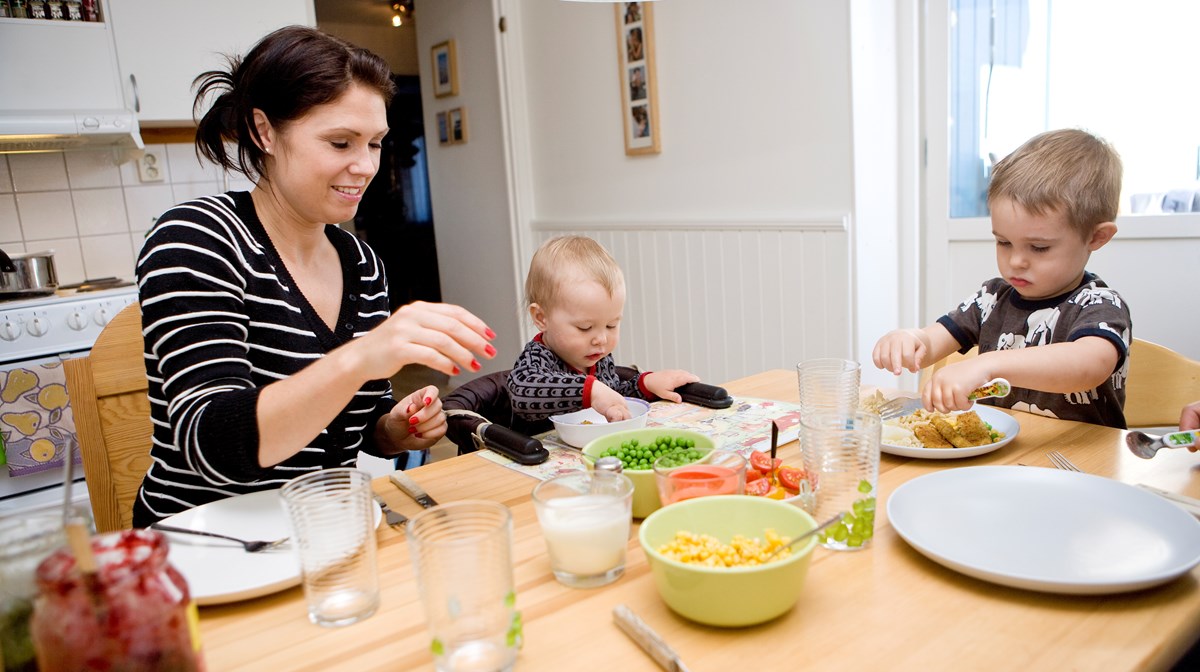 En mamma med två mindre barn sitter vid köksbordet. På bordet finns kålar med grönsaker.
