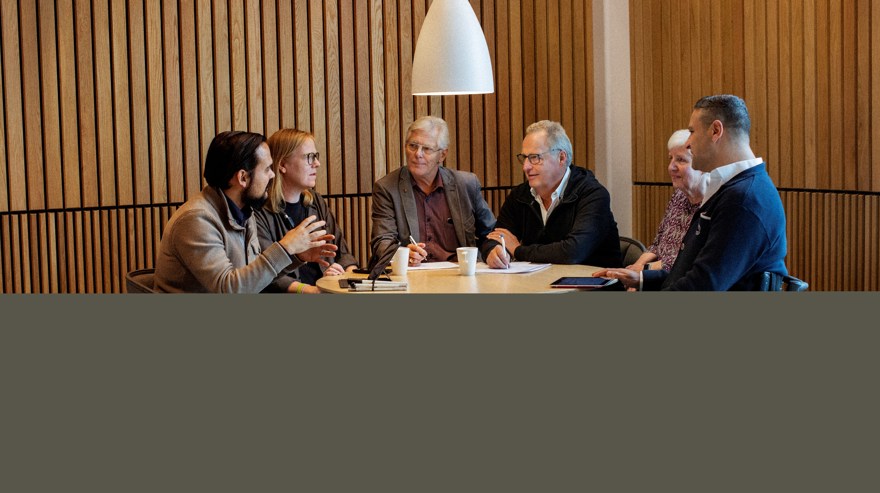 Sex personer sitter runt ett bord och samtalar. En av personerna sitter i rullstol.