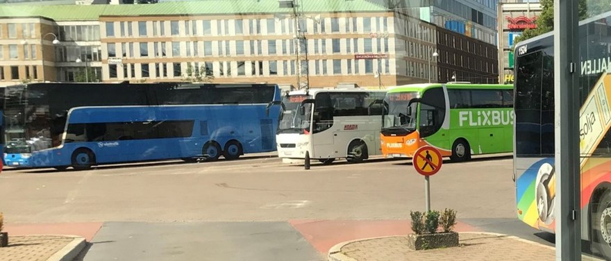 Fotografi genom glasrutan från Nils Ericson-terminalen i Göteborg. Utanför terminalen står parkerade bussar från olika bussföretag. Det är soligt sommarväder och köpcentret Nordstan syns i bakgrunden.