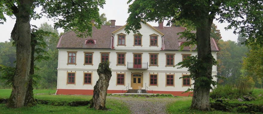 En vit byggnad med röda detaljer och mörkt tak står bakom träd.