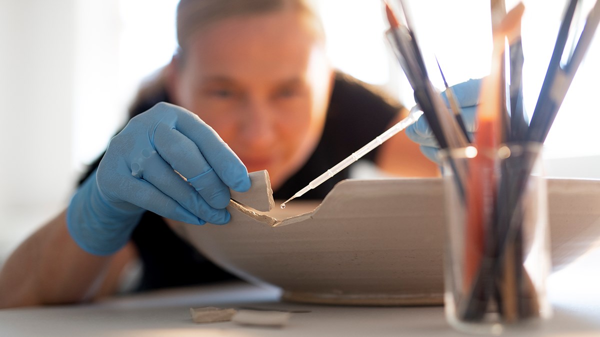 En konservator limmar försiktigt fast en skärva på ett trasigt keramikfat.