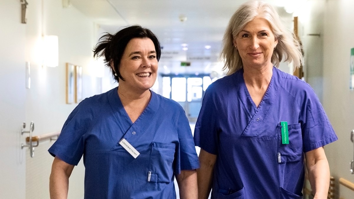 Två kvinnor i vårdklädsel går i en korridor i vårdmiljö