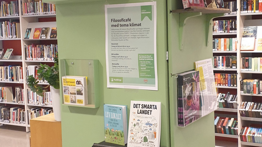 En ljusgrön hög möbel med fyra sidor står mitt i biblioteksrummet. På möbelns sidor finns hyllor och fack för böcker, broschyrer och annat. Mitt på den finns en plansch med rubriken Filosoficafé med tema klimat..