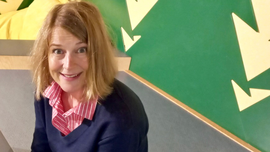 Hanna har rakt mörkblont hår till axlarna. Hon är klädd i en mörkblå v-ringad tröja och bär en röd- och vitrandig skjorta under den. Väggen bakom henne är målad i färgglada mönster.