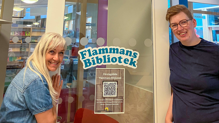 Towe Lindblom och Christoffer Svensson står på varsin sida om en glasdörr. På dörren finns texten "Flammans bibliotek" i blå bokstäver. Under den finns en lapp med texten "Förslagslåda Flammans bibliotek" och en QR-kod. Towe har långt ljust hår med lugg och är klädd i en jeansblus. Christoffer har kortklippt blont hår, bär glasögon och är klädd i en blå t-shirt. Båda två ler mot fotografen.