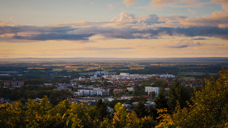 Skövde från berget Billingen. Nere i en dal ligger Skövde, vita höghus, höga skorstenar, röda hustak utgör staden. Bortom Skövde ser man långt ut över bygden, åkrar och skogspartier omvartannat, landskapet är platt så man ser långt.