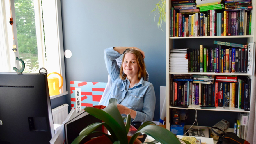 Camilla Eriksson sitter i sitt arbetsrum på biblioteket. Väggarna är ljusblå, bakom henne finns en bokhylla och på skrivbordet står en dataskärm och en krukväxt.