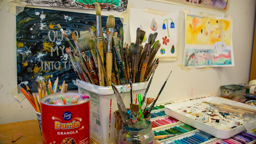 Penslar i en plastbytta, pennor i en annan. På bordet ligger färgkritor i rader och på väggen hänger målningar och teckningar.