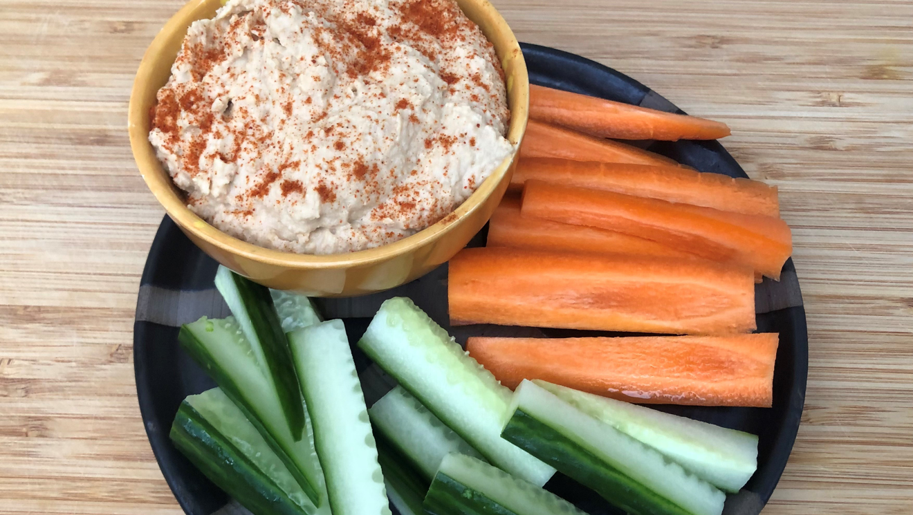 Hummus i en gul skål med stavar av morot och gurka på en svart tallrik