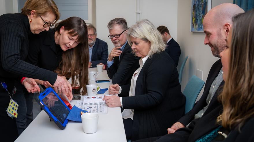 Ministern, en kvinna med ljust hår, tittar på när två medarbetare från Dart visar och pekar på kommunikationshjälpmedel