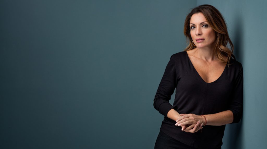 Alexandra Pascalidou har mörkt långt hår och stora bruna ögon. Hon är klädd i en svart tunn v-ringad tröja och mörka byxor. Bilden är tagen i en fotostudio, bakgrunden är duvblå, Alexandra lutar sig mot en vägg och tittar allvarligt in i kameran.