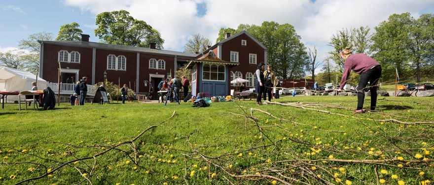 En röd byggnad med vita detaljer, flera personer utför olika aktiviteter på gräsmattan framför, kvistar ligger på gräsmattan