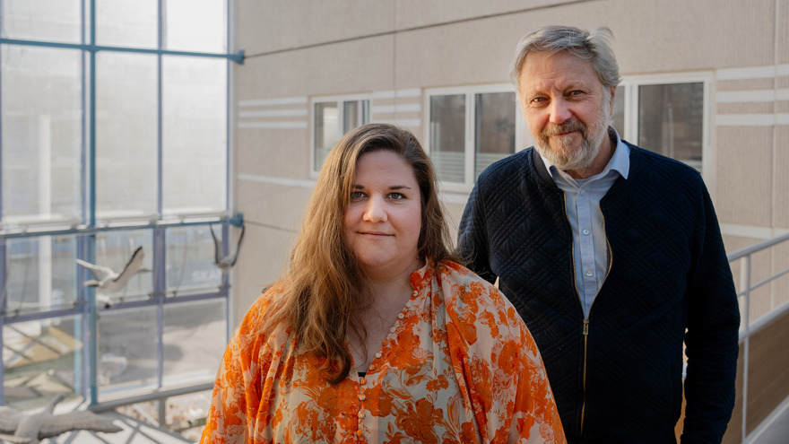 Frida Närman och Ricky Andreis står inomhus i någon slags ljusgård. Stora fönster och en hus vägg syns i bakgrunden. Frida har långt brunt hår och bär en mönstrad orange blus. Ricky har grått kortklippt hår och helskägg, han är klädd i ljusblå skjorta och en mörk cardigan med dragkedja.