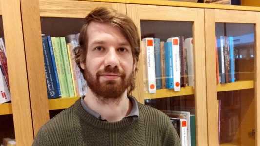 David Reichel har brunt hår och helskägg. Han bär en grön stickad tröja och står framför ett bokskåp fyllt med böcker.