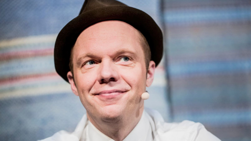 Olof Wretling är i medelåldern. Han har blå ögon och ljus hy, är klädd i en vit skjorta som är knäppt ända upp och på sitt huvud har han en brun hatt med korta brätten. Han har en liten mikrofon som är osynligt fäst bakom örat, han ler och hans blick är vänd åt höger i bild.