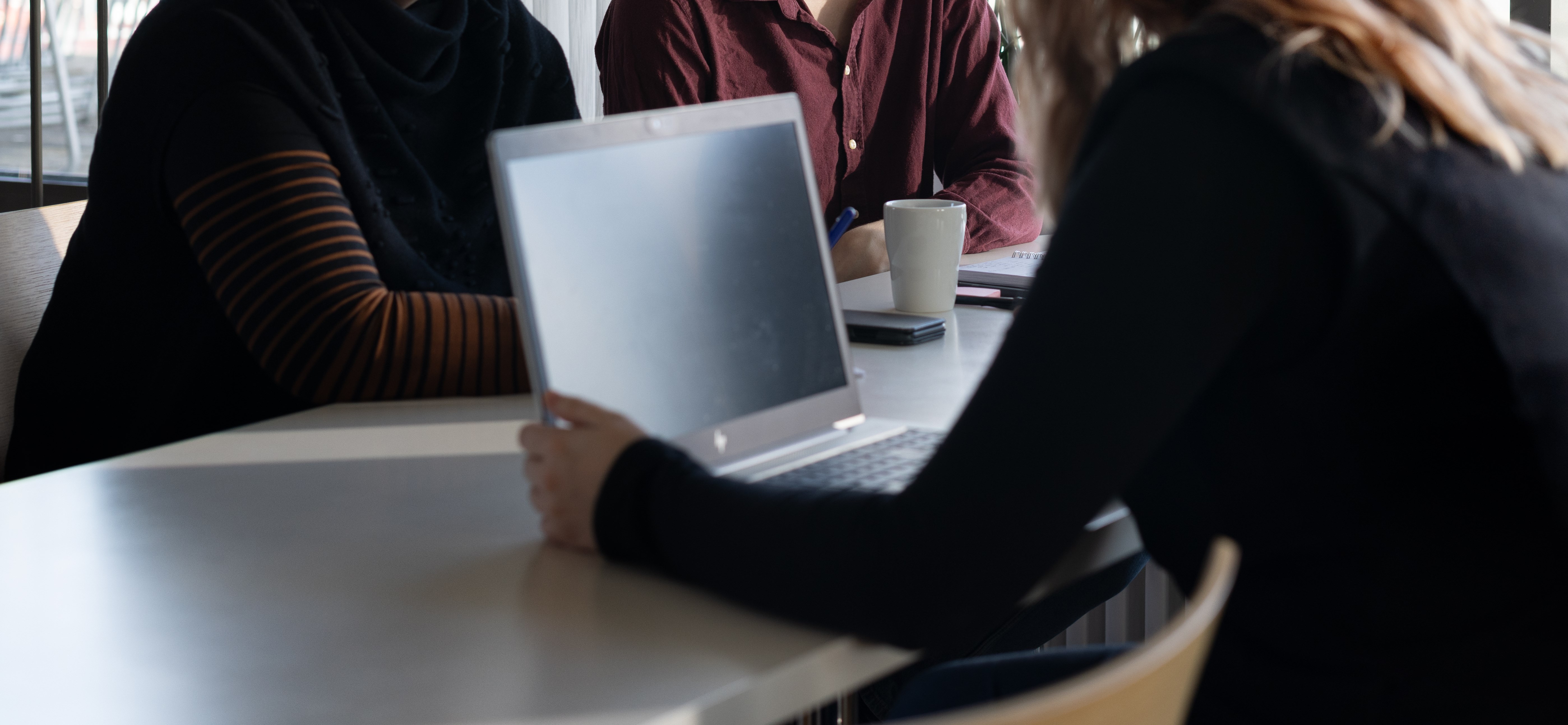 En bärbar dator på ett bord. Tre personer man endast ser överkropp på sitter runt bordet och datorn.