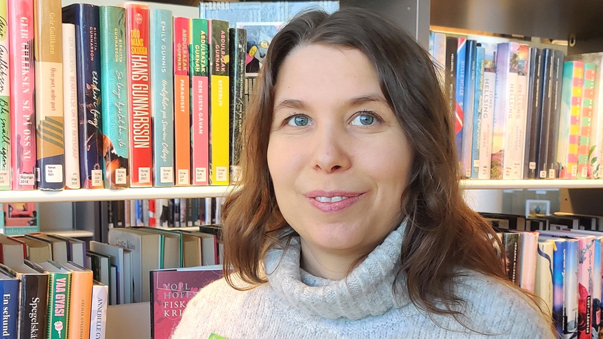 Ellen Ekbom har ljusbrunt långt hår och blå ögon. Hon står framför en bokhylla och ler mot kameran. Hon är klädd i en vit stickad tröja med polokrage.