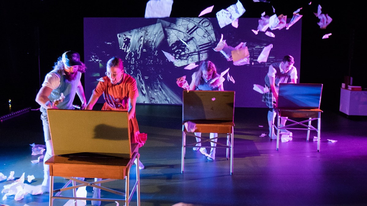 Teaterföreställning som visar personer vid skolbänkar och papper som flyger runt i luften.