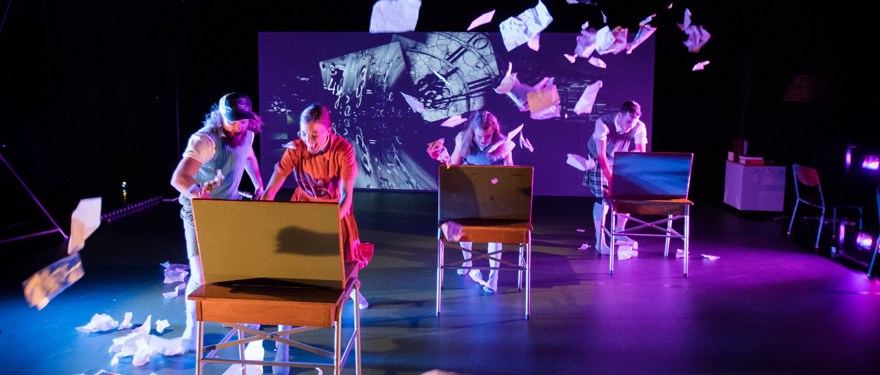 Teaterföreställning som visar personer vid skolbänkar och papper som flyger runt i luften.