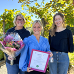 Med en bakgrund av färggranna lövträd står tre kvinnliga medarbetare från Sjukhusen i väster. Tillsammans med en blombukett håller personen i mitten i ett certifikat.