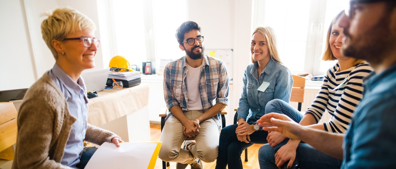 Helbild på fem personer som sitter på stolar i en cirkel. En av personerna verkar vara terapeut. Gruppen samtalar och ser glada ut.