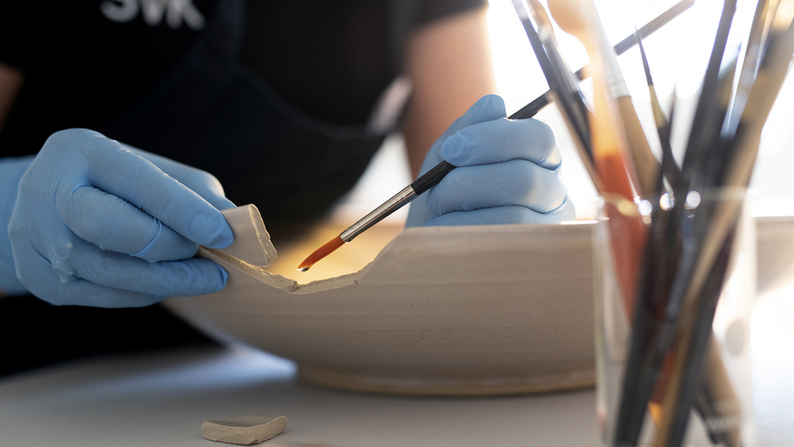 En person med ljusblå handskar håller en skärva som försiktigt limmas fast mot ett stort fat.
