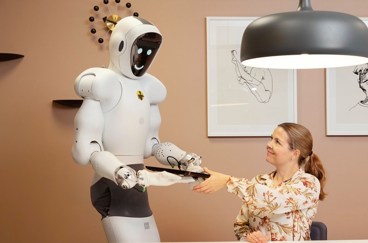 Vit, humaniod robot med runda former räcker laptop till sittande kvinna. 